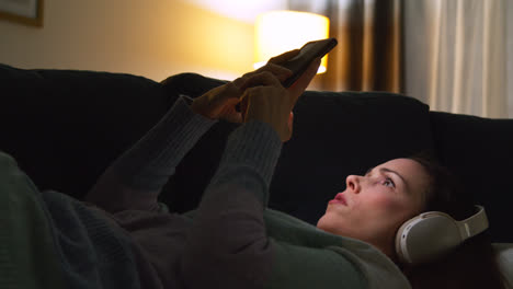 Mujer-Con-Auriculares-Inalámbricos-Tumbada-En-El-Sofá-De-Casa-Por-La-Noche-Transmitiendo-O-Mirando-Contenido-En-Línea-En-El-Teléfono-Móvil-2